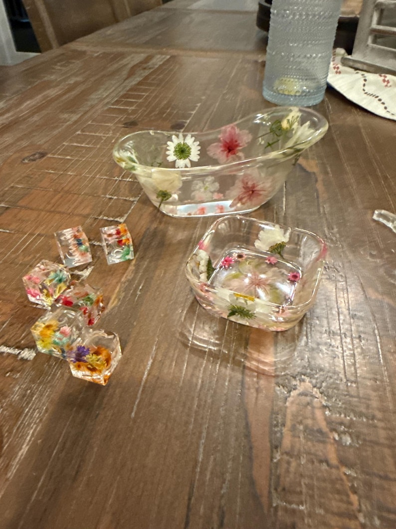 May include: Clear resin decorative pieces with embedded dried flowers. The image shows a small bathtub-shaped dish, a square dish, and several small, cube-shaped resin pieces. The flowers are various colors, including white, pink, and yellow, set against a clear background.