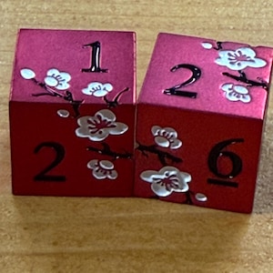 May include: Two red dice with white floral designs and black numbers. One die shows the number 1, and the other shows the numbers 2 and 6. The dice have a metallic sheen and are likely used for tabletop games.