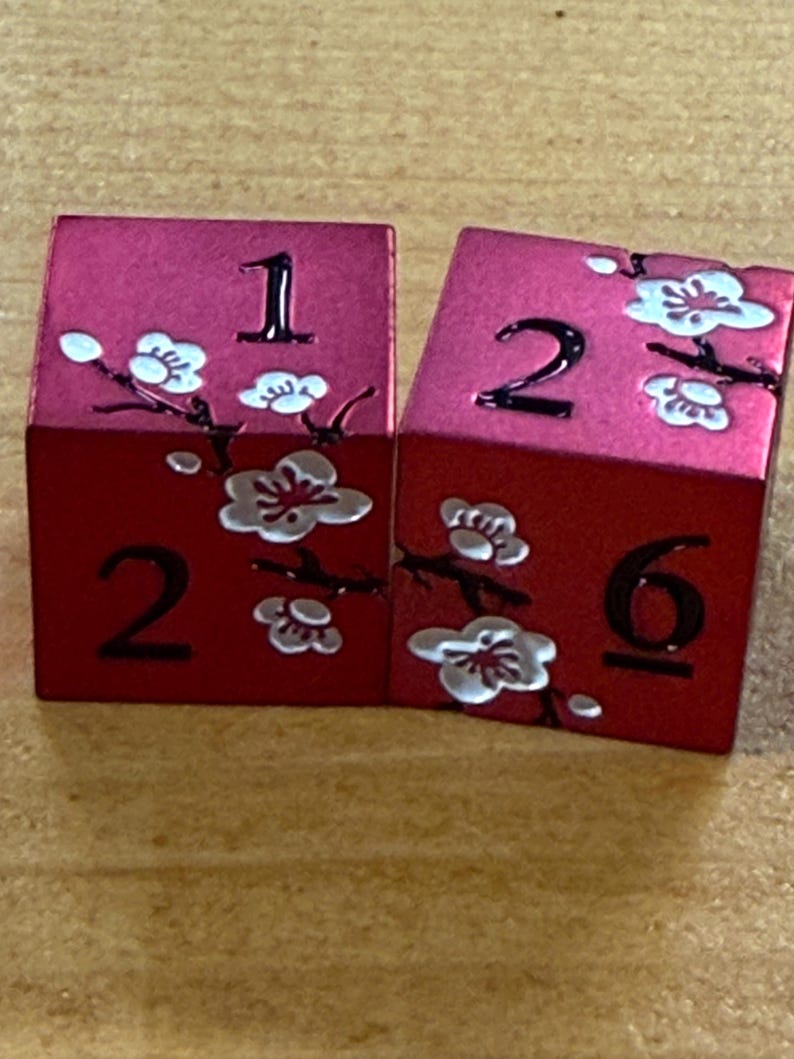 May include: Two red dice with white floral designs and black numbers. One die shows the number 1, and the other shows the numbers 2 and 6. The dice have a metallic sheen and are likely used for tabletop games.