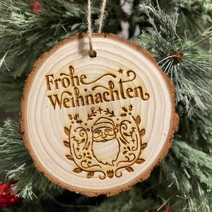 May include: A wooden Christmas bauble with a rustic, natural wood finish. It features a laser-engraved design of Santa Claus with a beard and a wreath of holly and leaves. The bauble also has the text "Frohe Weihnachten" which is German for "Merry Christmas".
