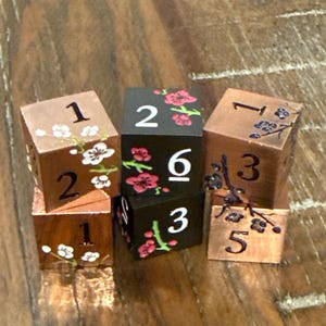 May include: Six wooden dice with floral designs and numbers. The dice are copper and black, with white and red floral accents. Numbers are painted in white. The dice are arranged in a small pile.