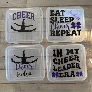 May include: Four clear plastic storage boxes with cheerleading themed designs. The boxes have black and purple text and images. The text on the boxes reads "Cheer", "Eat Sleep Cheer Repeat", "In My Cheerleader Era", and "Cheer Jaidyn".