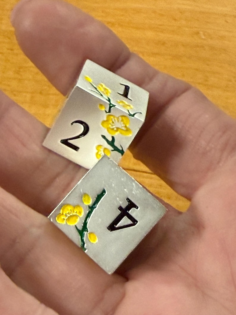 May include: Two silver dice with black numbers and yellow floral designs. One die shows the numbers 1 and 2, while the other shows the number 4. The dice are held in a hand.