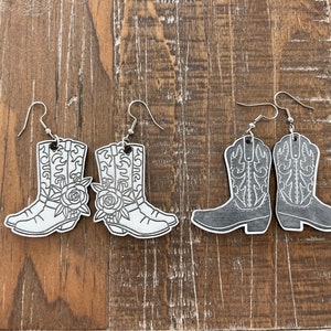 Könnte beinhalten: Zwei Paar Hängeohrringe mit einem Cowboy-Stiefel-Design. Ein Paar ist weiß mit schwarzen Umrissen und einem Blumendesign, das andere Paar ist schwarz mit weißen Umrissen.