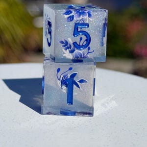 May include: Two clear resin dice, one stacked on top of the other. The dice feature blue floral designs and blue numbers. The top die shows the number 5, while the bottom die shows the number 1. The dice are set against a blurred background.