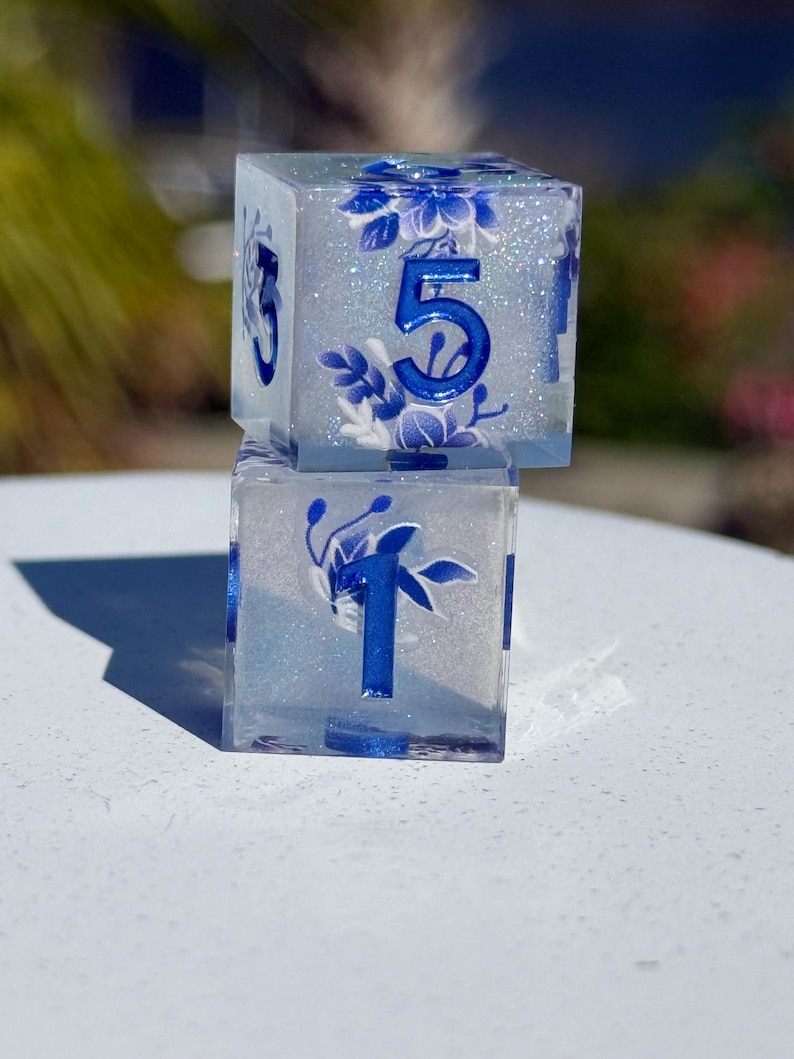 May include: Two clear resin dice, one stacked on top of the other. The dice feature blue floral designs and blue numbers. The top die shows the number 5, while the bottom die shows the number 1. The dice are set against a blurred background.