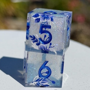 May include: Two clear, square dice with blue floral designs and glitter. The dice have the numbers 5 and 6 in blue. The dice are stacked on top of each other.