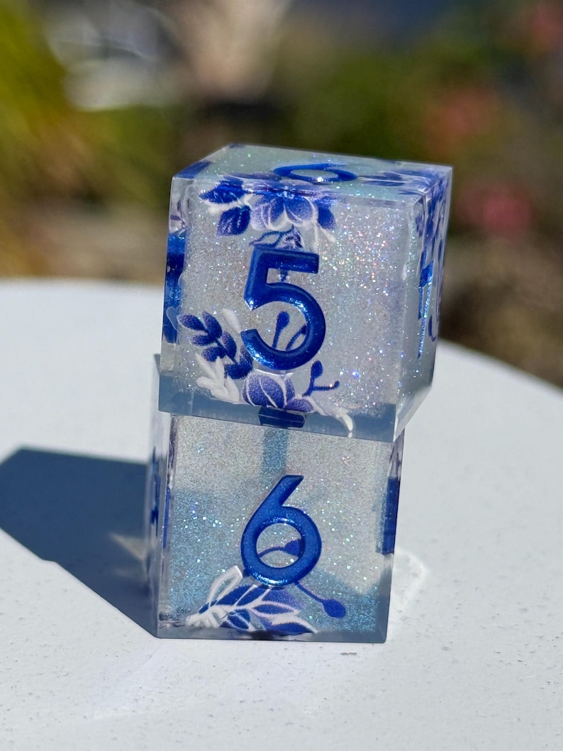 May include: Two clear, square dice with blue floral designs and glitter. The dice have the numbers 5 and 6 in blue. The dice are stacked on top of each other.