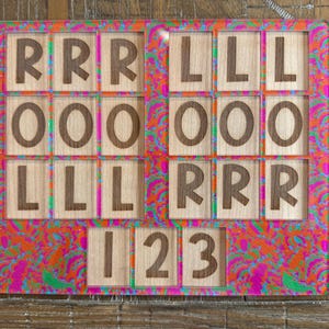May include: A rectangular board featuring a vibrant, multi-colored psychedelic pattern in shades of pink, orange, and green. The board contains multiple square wooden tiles engraved with the letters R, L, O, and the numbers 1, 2, and 3.