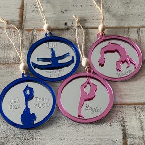 Könnte beinhalten: Vier runde, silberfarbene, spiegelnde Ornamente mit einer Turner-Silhouette in der Mitte. Die Ornamente sind blau, rosa und zwei sind silber. Die Ornamente haben Text darauf, darunter "Liv", "Dakota Spirit 2024-25", "Team YUGO", "USA 2024", "Baylor" und "Rowan".
