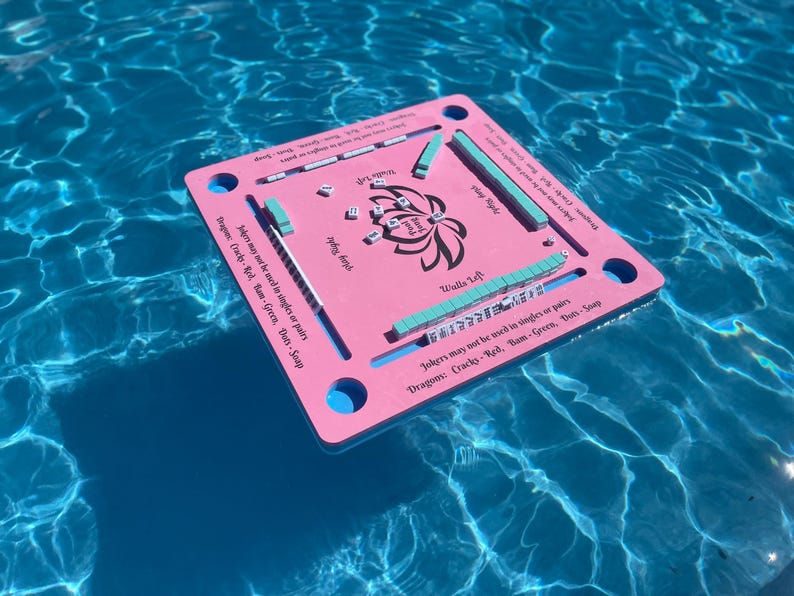Puede incluir: Un tablero de juego flotante de color rosa y azul con las palabras "Jokers may not be used in singles or pairs" y "Dragon: Cracks-Red, Bam-Green, Dots-Saap" impresas en &eacute;l. El tablero tiene un dise&ntilde;o de flor de loto en el centro y flota en una piscina azul.