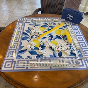 May include: Mahjong set on a round wooden table. The game board features a blue and white floral design with yellow accents. A blue case with "Mahj Tiles" printed on it sits nearby. The set includes tiles and yellow tile racks.