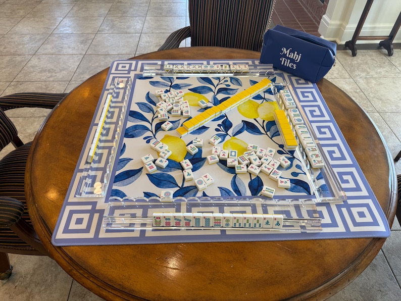 May include: Mahjong set on a round wooden table. The game board features a blue and white floral design with yellow accents. A blue case with "Mahj Tiles" printed on it sits nearby. The set includes tiles and yellow tile racks.