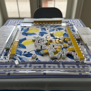 May include: A Mahjong set with yellow and white tiles scattered on a table. The tiles are arranged on a clear acrylic playing surface. The table is covered with a blue and white patterned tablecloth, with a lemon design.