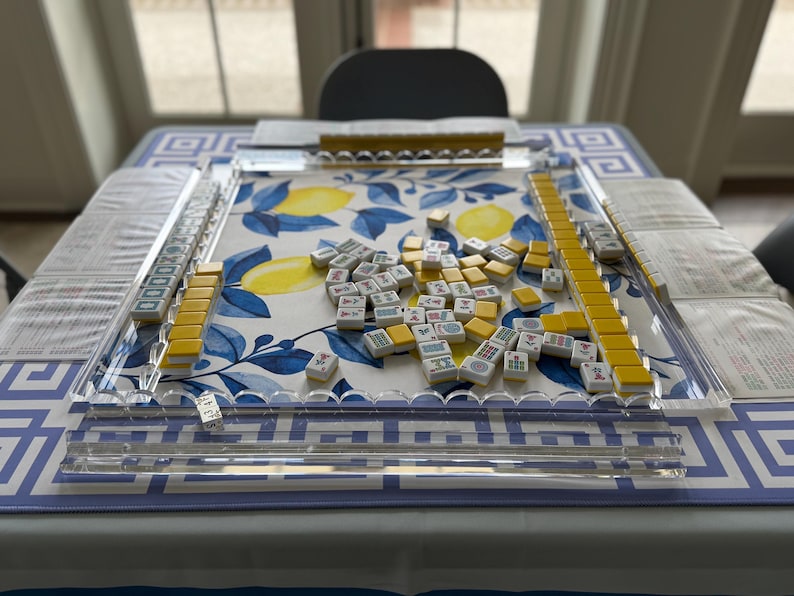 May include: A Mahjong set with yellow and white tiles scattered on a table. The tiles are arranged on a clear acrylic playing surface. The table is covered with a blue and white patterned tablecloth, with a lemon design.