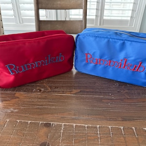 May include: Two rectangular cosmetic bags. One is red with blue embroidered text, and the other is blue with red embroidered text. Both bags have a zipper closure and are sitting on a wooden surface. The text on both bags reads "Rummikub".