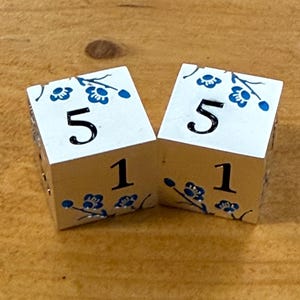 May include: Two white dice with blue floral designs and the numbers 5 and 1. The dice are on a wooden surface. The blue floral design is on the top and bottom of the dice.