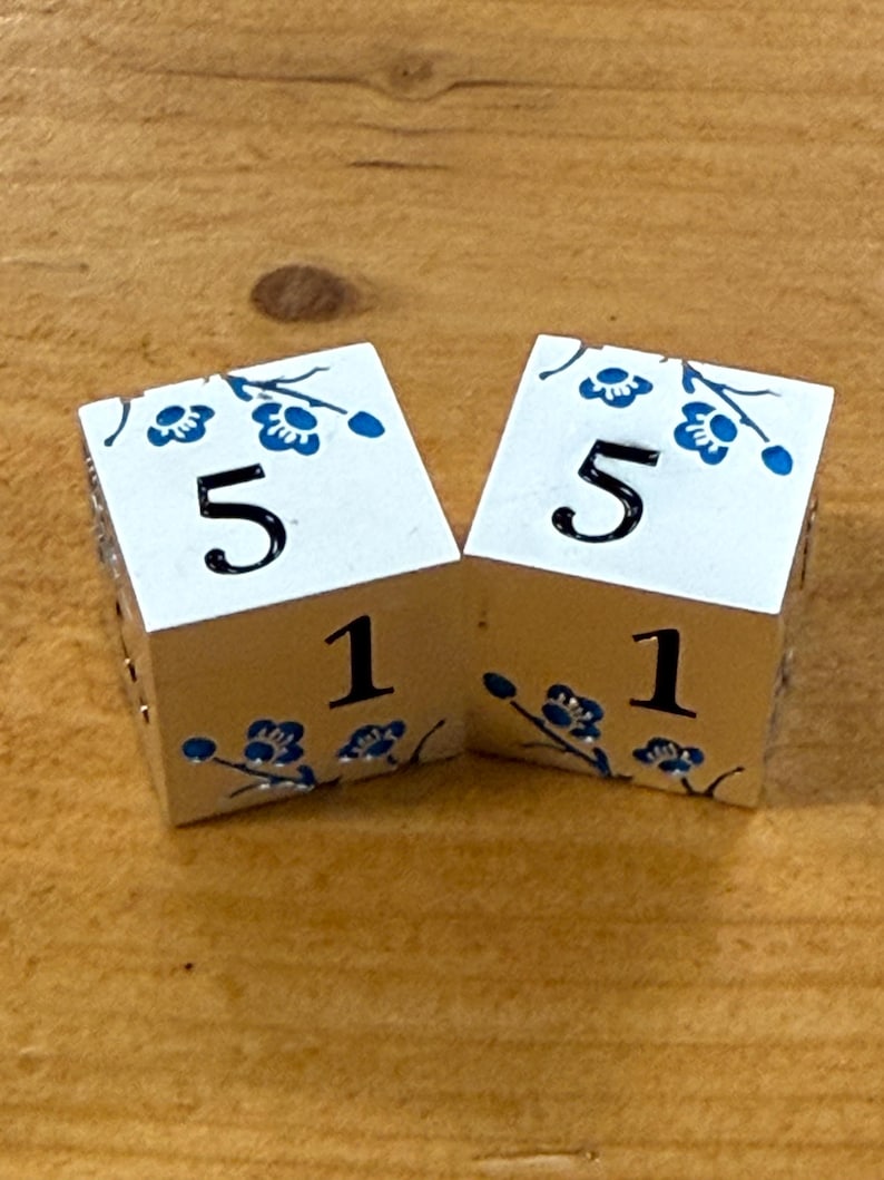 May include: Two white dice with blue floral designs and the numbers 5 and 1. The dice are on a wooden surface. The blue floral design is on the top and bottom of the dice.