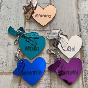 Puede incluir: Cinco llaveros en forma de corazón con superficies espejadas. Cada llavero presenta un par de pequeñas huellas de pies y las palabras "Mommy", "Mom", "Jad" y "Mommy" en letras plateadas. Los llaveros son de varios colores, incluyendo oro rosa, plata, azul verdoso, azul y morado.