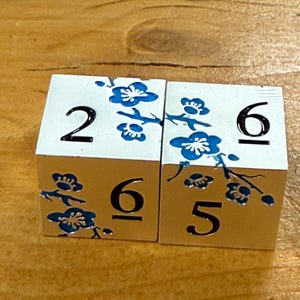 May include: Two white wooden blocks with black numbers and blue floral designs. One block shows the number 2, and the other shows the number 6. The blocks are on a wooden surface.