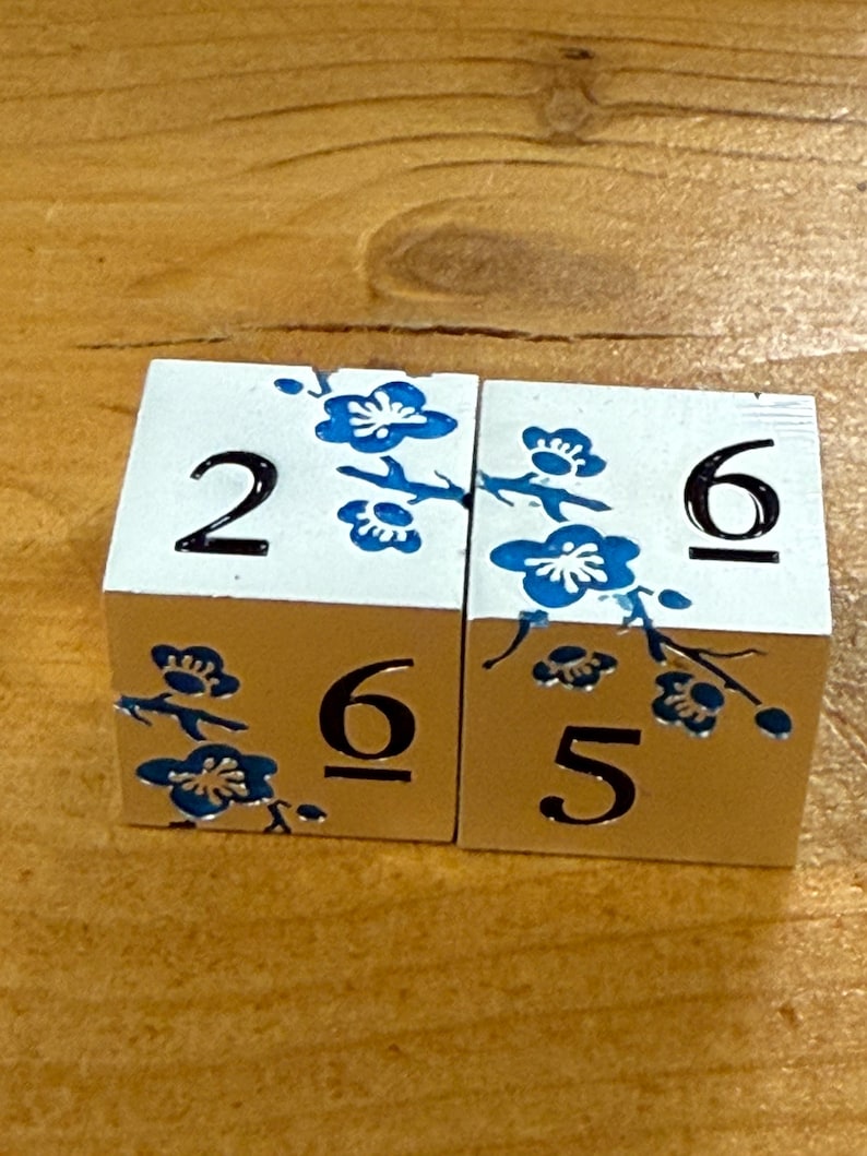May include: Two white wooden blocks with black numbers and blue floral designs. One block shows the number 2, and the other shows the number 6. The blocks are on a wooden surface.