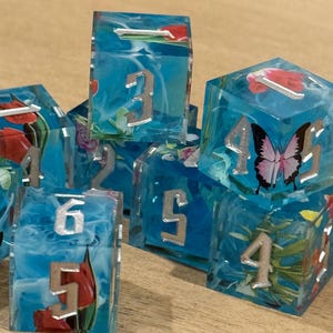 May include: A collection of handmade resin dice. Each die is a translucent blue with silver numbers and embedded floral and butterfly designs. The dice are arranged on a wooden surface.