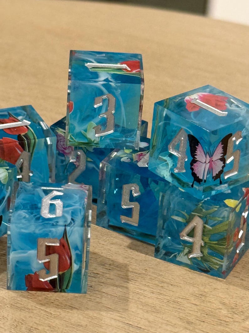 May include: A collection of handmade resin dice. Each die is a translucent blue with silver numbers and embedded floral and butterfly designs. The dice are arranged on a wooden surface.