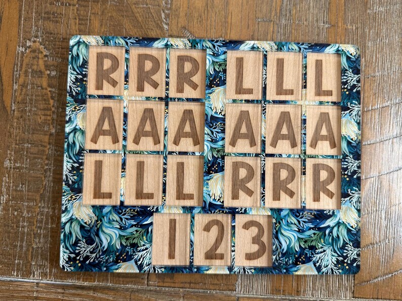 May include: A rectangular wooden puzzle board featuring a dark blue and teal floral pattern border. The board contains square wooden tiles engraved with the letters R, L, and A, and the numbers 1, 2, and 3, arranged in a grid-like layout.