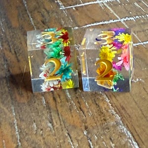 May include: Two clear resin dice with gold numbers and colorful dried flowers inside. The dice are sitting on a wooden surface. The numbers on the dice are 2 and 3.
