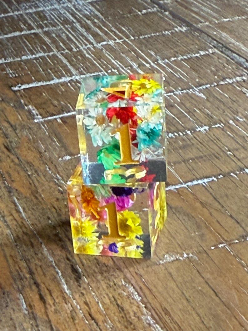 May include: Two clear resin cubes, each containing colorful dried flowers and a gold numeral "1". The flowers include white daisies, yellow, red, and purple blooms. The cubes are stacked on a wooden surface.