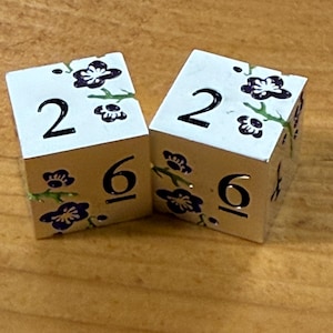 May include: Two white dice with silver edges, each displaying the number 2 on top and 6 on the side. The dice are decorated with a floral design in purple and green. The dice are on a wooden surface.