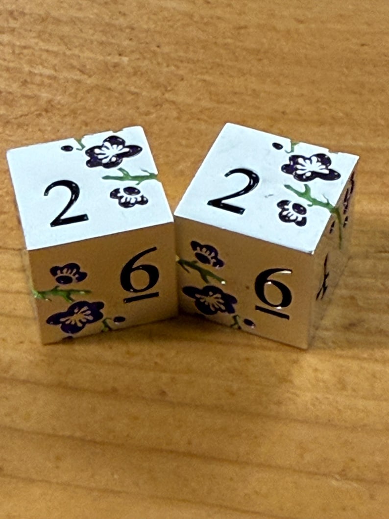 May include: Two white dice with silver edges, each displaying the number 2 on top and 6 on the side. The dice are decorated with a floral design in purple and green. The dice are on a wooden surface.