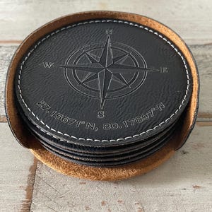 May include: A set of black leather coasters with a compass rose design and white stitching. The coasters are in a brown holder. Coordinates 27.15671°N, 80.17897°W are printed on the coasters.