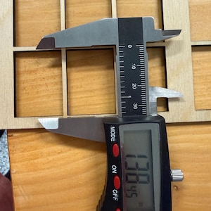 May include: A light-colored wooden grid panel with square cutouts being measured by a digital caliper. The caliper display shows a measurement of 1.3845 inches, indicating the width of one of the square openings in the wooden grid structure.