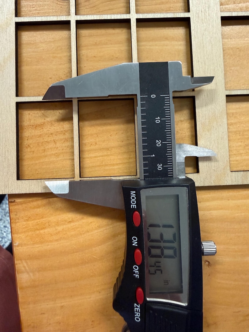 May include: A light-colored wooden grid panel with square cutouts being measured by a digital caliper. The caliper display shows a measurement of 1.3845 inches, indicating the width of one of the square openings in the wooden grid structure.