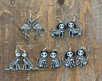 Puede incluir: Un conjunto de ocho pendientes de esqueleto con un flamenco, gatos y perros. Los pendientes están hechos de acrílico blanco y negro y tienen ganchos plateados.
