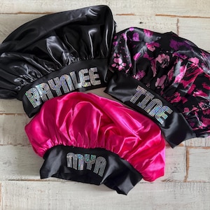 May include: Three satin sleep caps with black trim and rhinestone lettering. The caps are pink, black, and black with a pink and purple floral print. The names "Mya", "Brylee", and "Time" are spelled out on the caps.