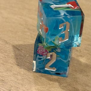 May include: Two dice made of clear resin with a blue and white swirl design. The dice have metallic numbers and floral accents. One die shows the number 3, and the other shows the number 2.