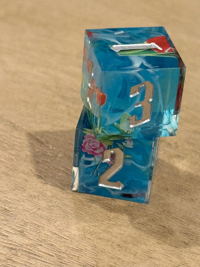 May include: Two dice made of clear resin with a blue and white swirl design. The dice have metallic numbers and floral accents. One die shows the number 3, and the other shows the number 2.