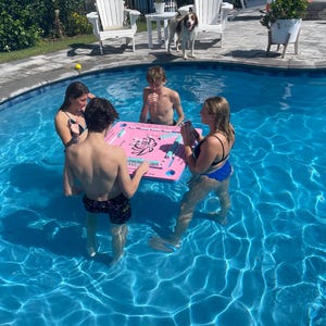 Puede incluir: Cuatro personas est&aacute;n jugando a los domin&oacute;s en una mesa flotante rosa en una piscina. La mesa tiene un dise&ntilde;o floral y los domin&oacute;s son visibles. La piscina es azul y el agua est&aacute; clara.