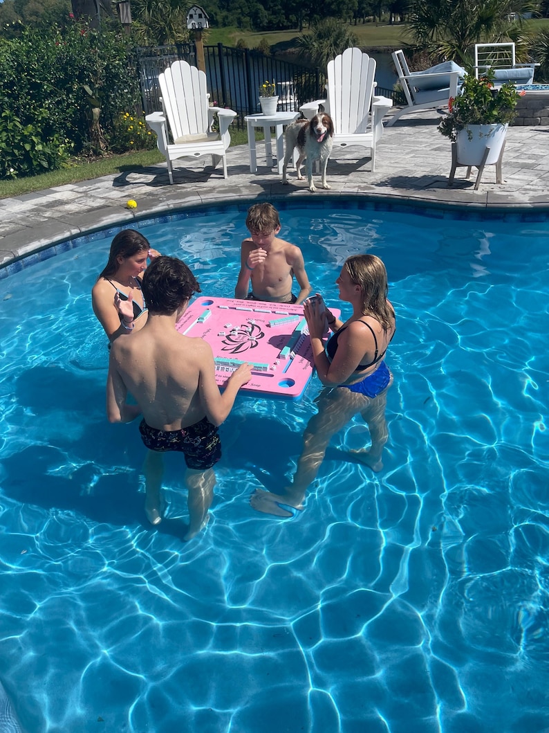 Puede incluir: Cuatro personas est&aacute;n jugando a los domin&oacute;s en una mesa flotante rosa en una piscina. La mesa tiene un dise&ntilde;o floral y los domin&oacute;s son visibles. La piscina es azul y el agua est&aacute; clara.