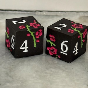 May include: Two black dice with white numbers and pink floral accents. The dice show the numbers 2, 4, and 6. The pink flowers have green stems.