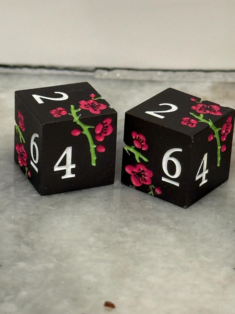 May include: Two black dice with white numbers and pink floral accents. The dice show the numbers 2, 4, and 6. The pink flowers have green stems.