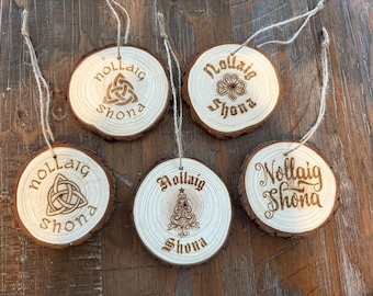 Könnte beinhalten: Sechs Weihnachtsbaumschmuck aus Holz mit Kordelaufhängern. Jeder Schmuck ist eine kreisförmige Holzscheibe mit natürlichem Rindenrand. Die Ornamente sind mit den Worten "Nollaig Shona" und keltischen Designs graviert, darunter ein Kleeblatt und ein Weihnachtsbaum.