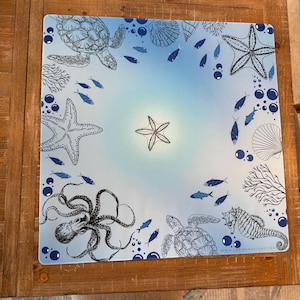 May include: Square placemat with a marine theme. The placemat features illustrations of sea creatures, including turtles, starfish, an octopus, and a seahorse, in shades of blue and white against a gradient blue background.