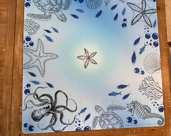 May include: Square placemat with a marine theme. The placemat features illustrations of sea creatures, including turtles, starfish, an octopus, and a seahorse, in shades of blue and white against a gradient blue background.