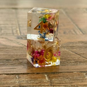 May include: Two clear resin dice with gold numbers and colorful dried flowers inside. One die shows the number 4, and the other shows the number 6. The dice are displayed on a wooden surface.