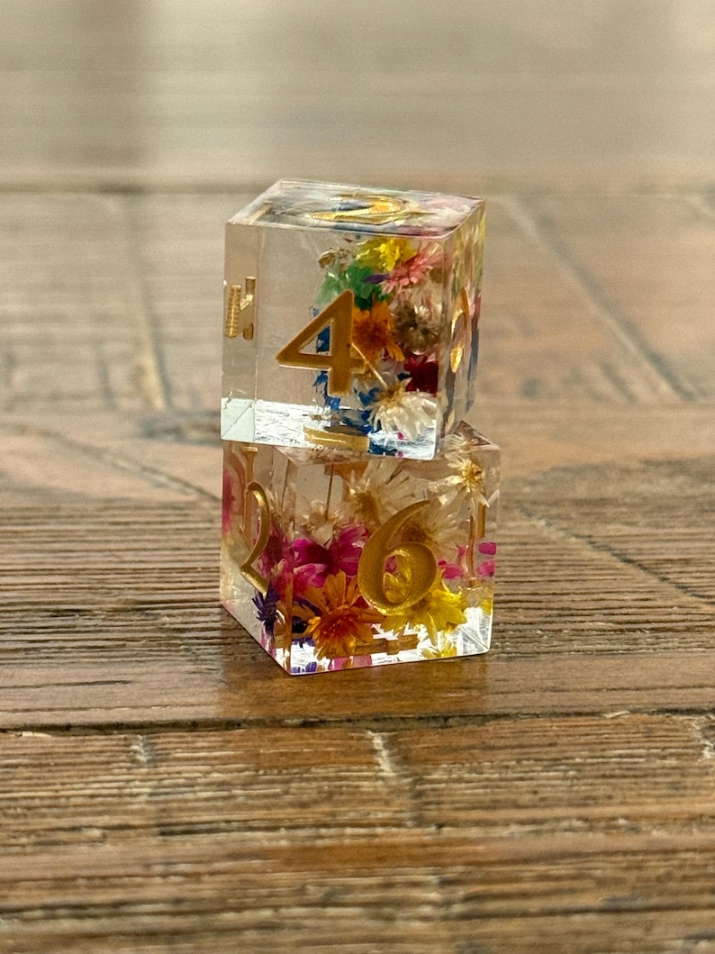 May include: Two clear resin dice with gold numbers and colorful dried flowers inside. One die shows the number 4, and the other shows the number 6. The dice are displayed on a wooden surface.