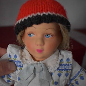 Könnte beinhalten: Eine Vintage-Puppe mit blauen Augen, blondem Haar und rosigen Wangen. Die Puppe trägt eine rot-schwarz-weiß gestrickte Mütze und eine weiße Bluse mit blauer Blumenstickerei. Eine hellblaue Schleife ziert die Bluse.