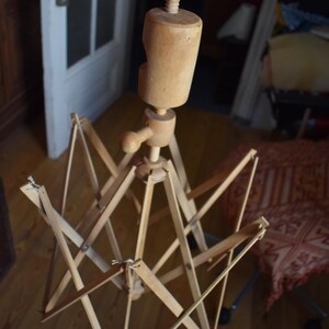 May include: A wooden yarn swift, a tool for winding yarn into a skein. The swift is made of light-colored wood and has an adjustable umbrella-like design. The wooden swift is held by a hand, showcasing its size and construction.